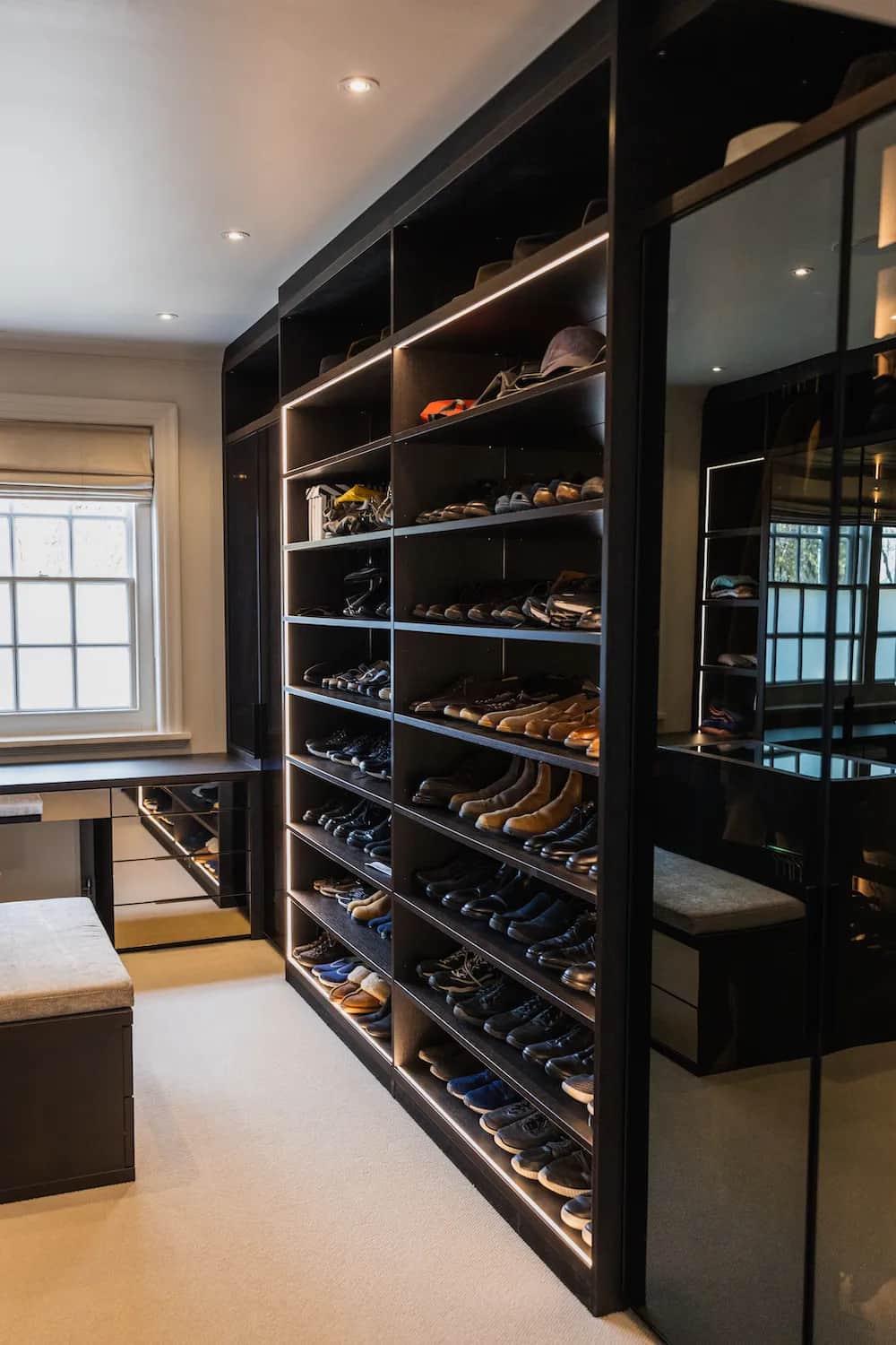 Dark walk-in wardrobe with lit shoe shelving, mirrored doors and a bench